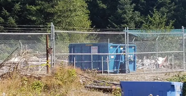 A blue trash can located behind a chain link fence, creating a contrast with the surrounding environment.